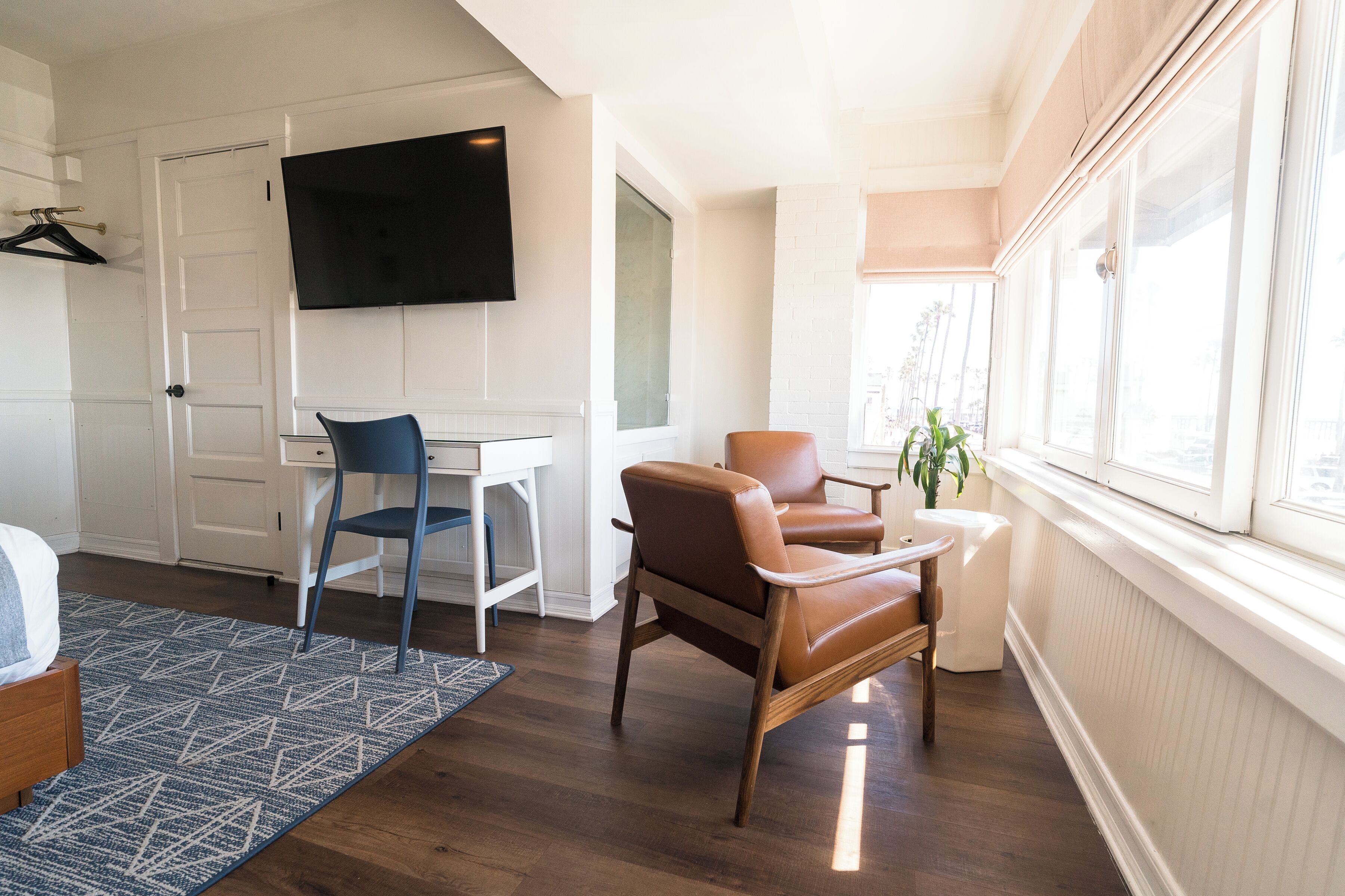 A modern hotel room with a flat-screen TV, a desk, a brown leather chair, a blue chair, and a large window with a view.