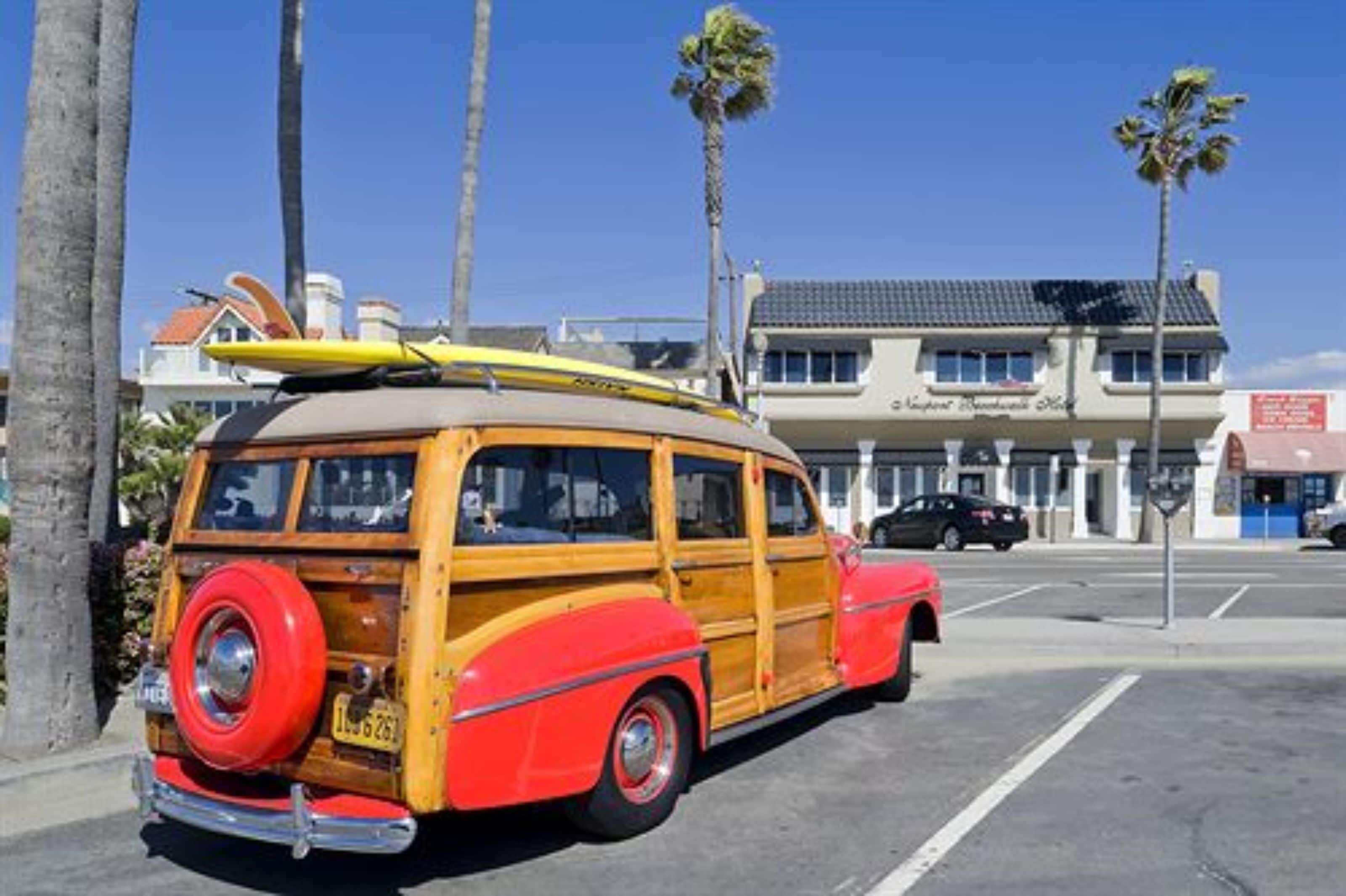 A vintage red and yellow station wagon parked in a lot with palm trees and a building in the background.
