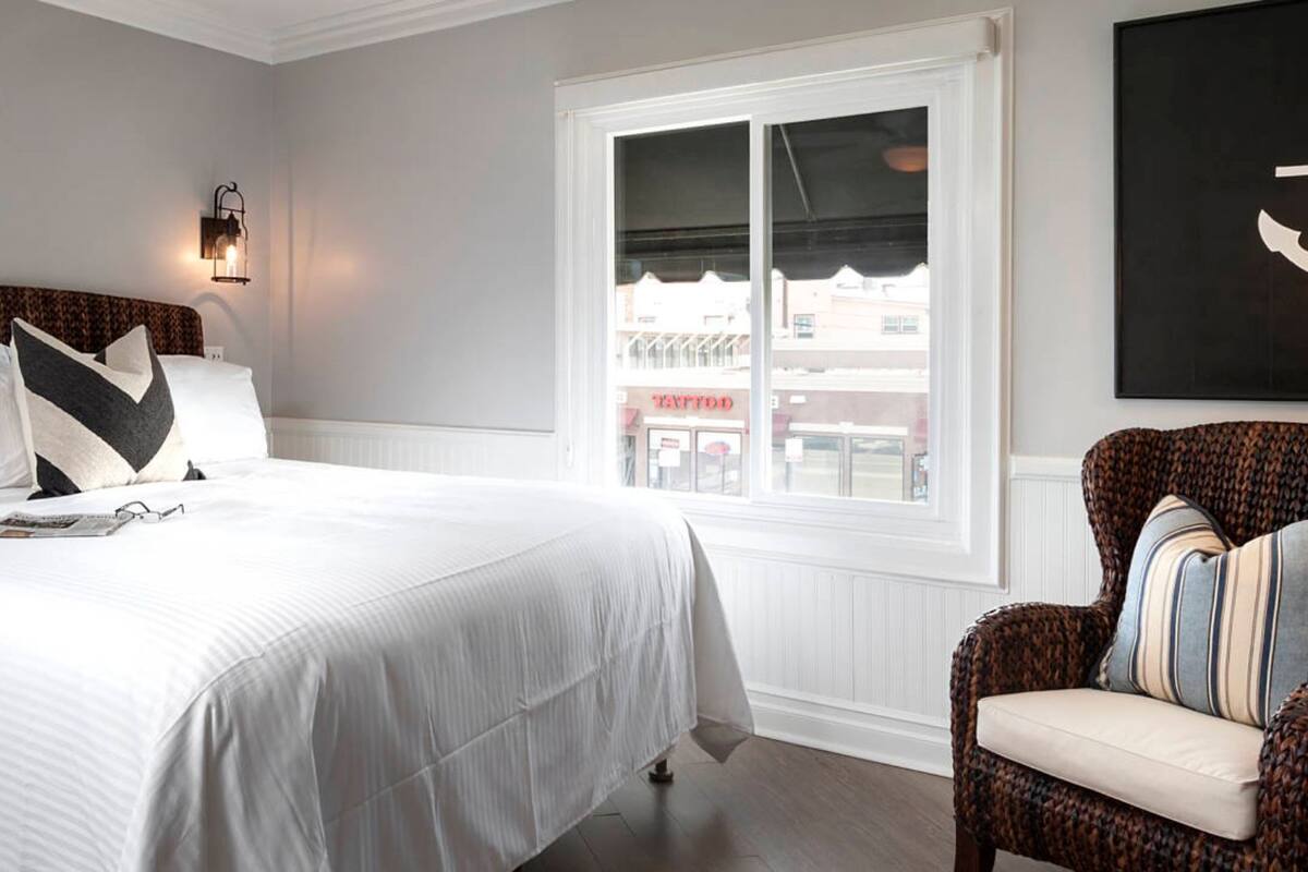 Een slaapkamer met een groot bed, een rieten stoel, een dressoir en een raam met uitzicht op een tattooshop.