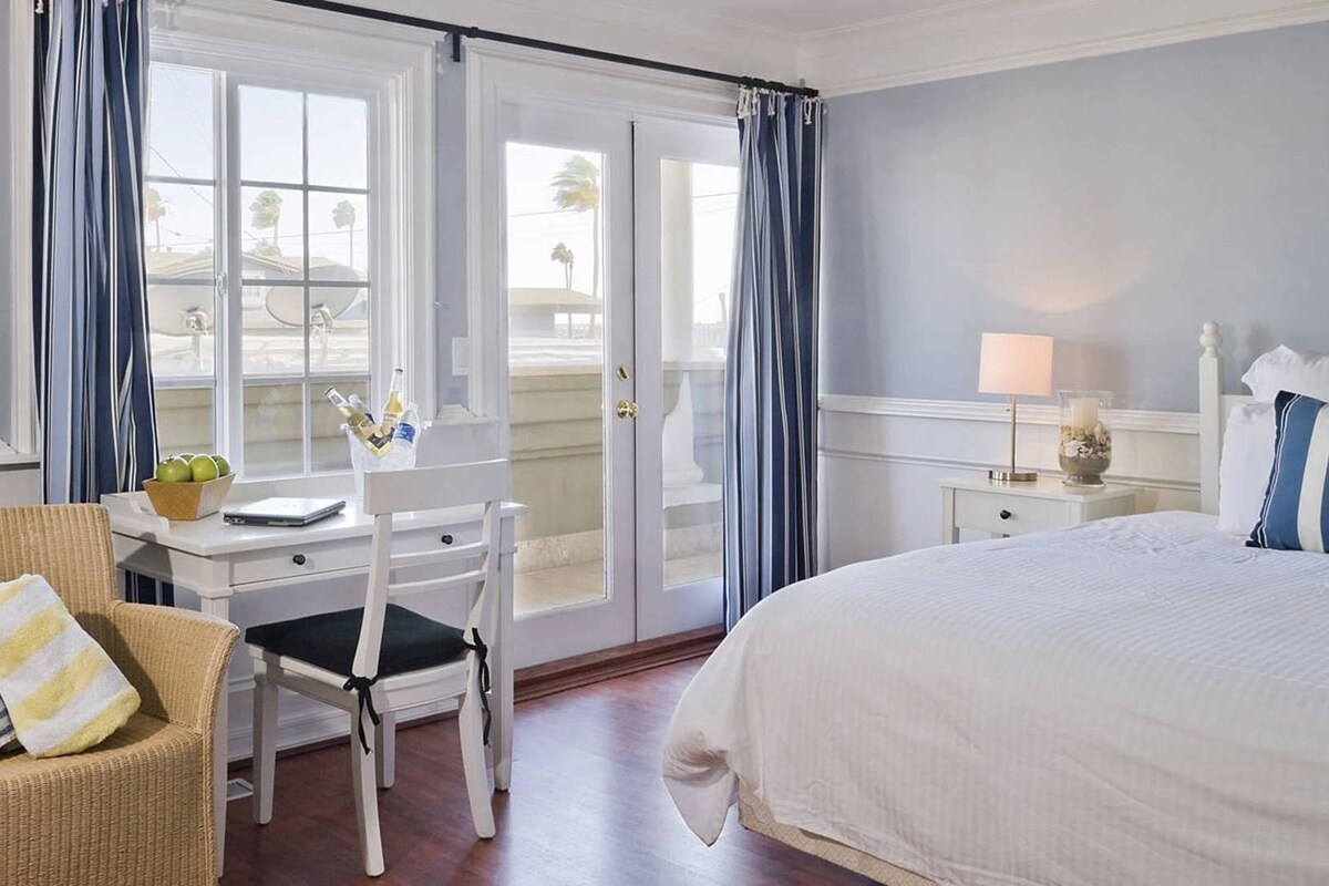A hotel room with a bed, a desk with a chair, a nightstand with a lamp, and a window with curtains.