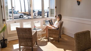 A room with wicker chairs, a small table, and a large window with a view of the beach and parked cars.