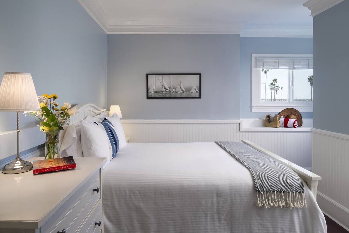 Een slaapkamer met een bed, nachtkastje, lamp en een raam met uitzicht op palmbomen.