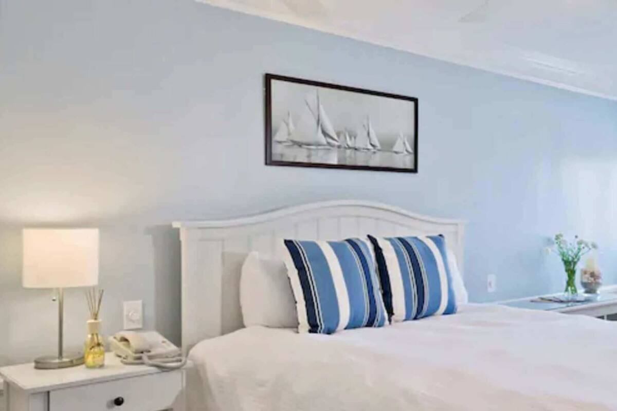 Hotelkamer met een wit bed, gestreepte kussens, een nachtkastje met een lamp en een ingelijst schilderij aan de muur.