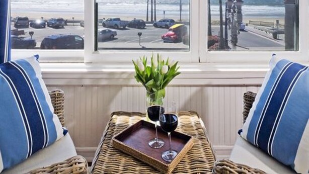 A room with a view of the beach, a wicker chair, two pillows, and a tray with two wine glasses.