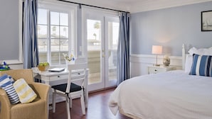 A hotel room with a bed, a desk with a chair, a window with curtains, a nightstand with a lamp, and a framed picture on the wall.