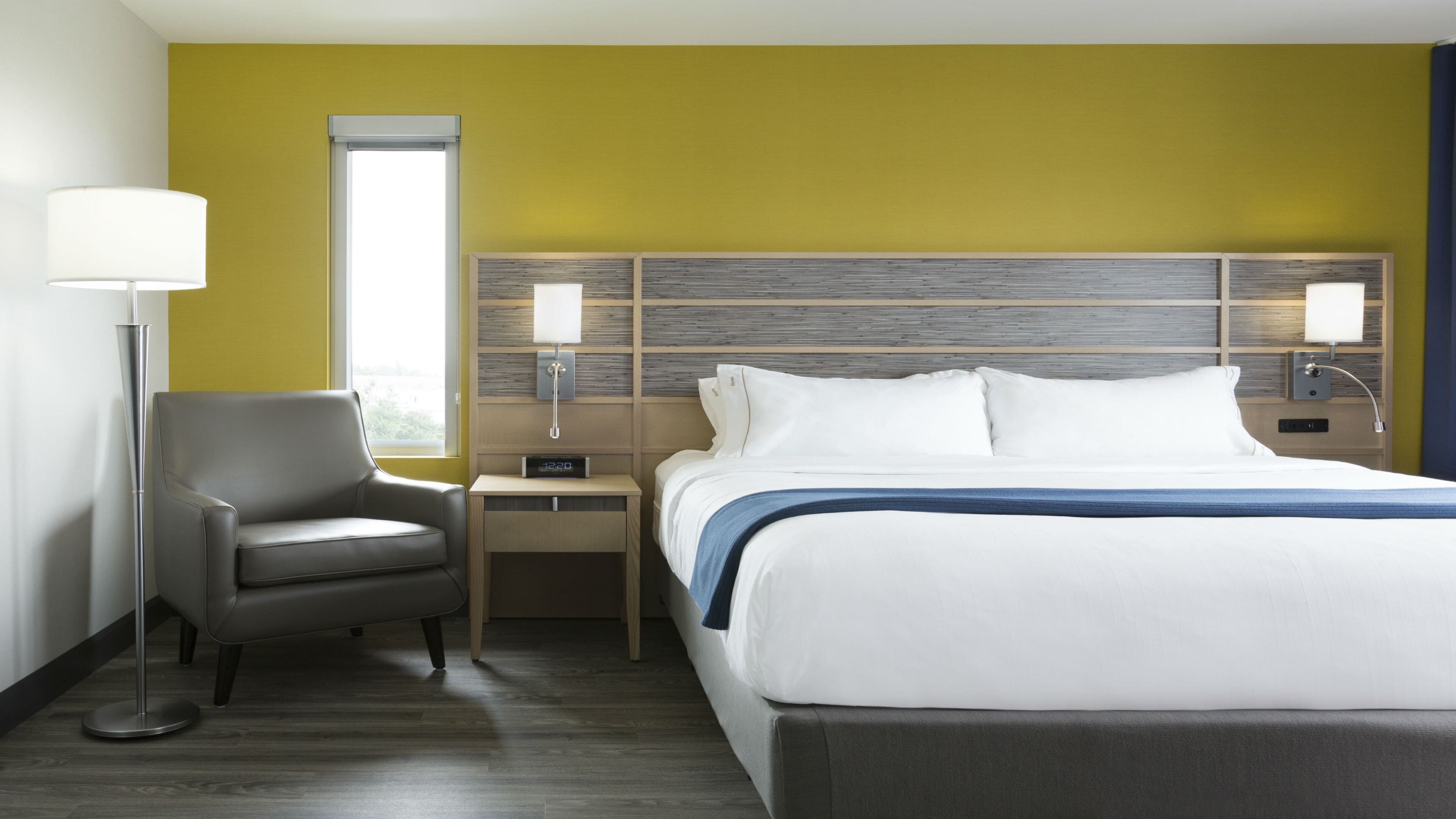 Une chambre d’hôtel avec un lit, une chaise, une table de chevet, une lampe et une fenêtre.