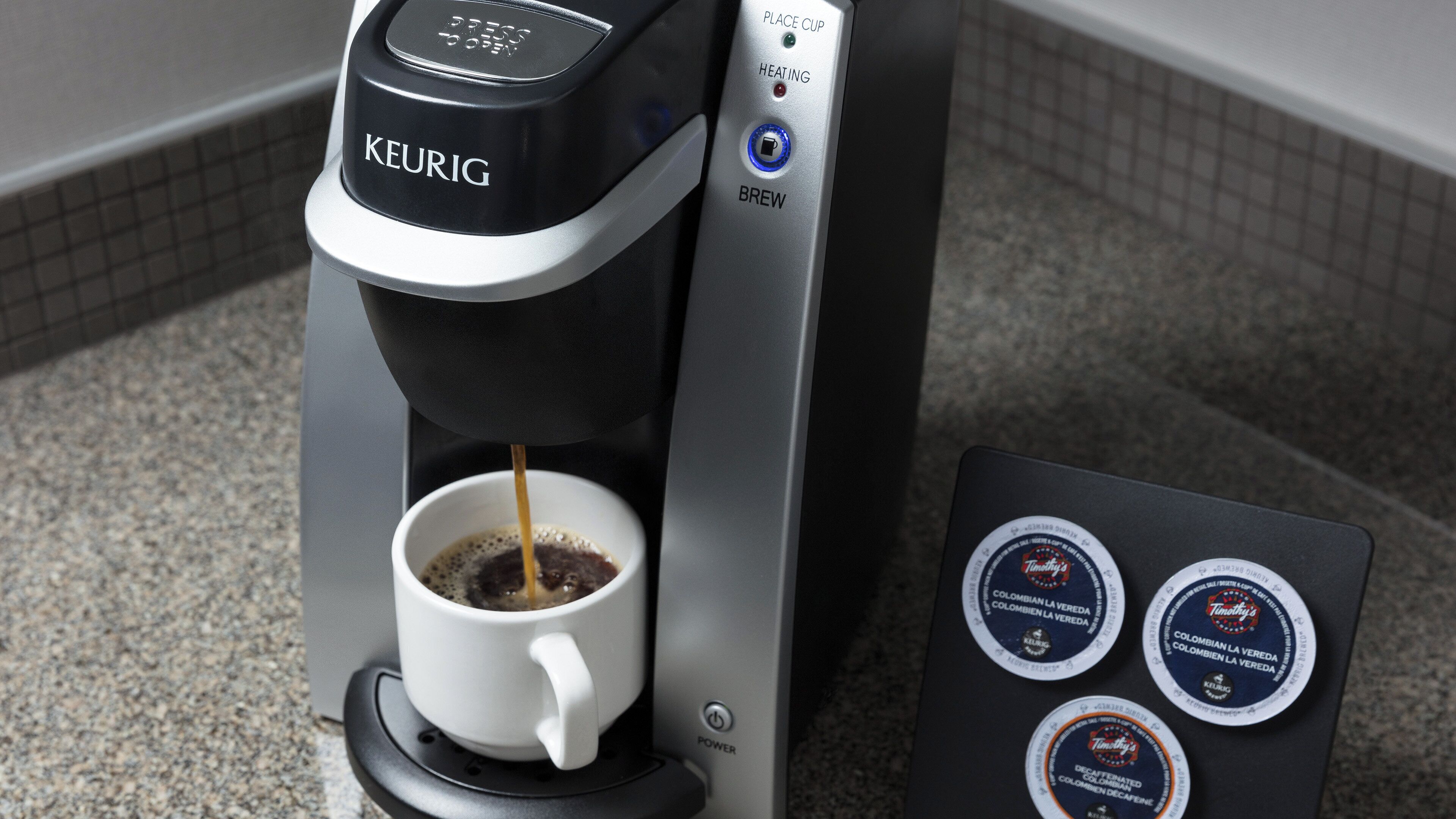 Una cafetera Keurig con una taza de café preparándose, un botón de encendido y dos cápsulas de café con la etiqueta 'Timothy's Colombian La Vereda'.