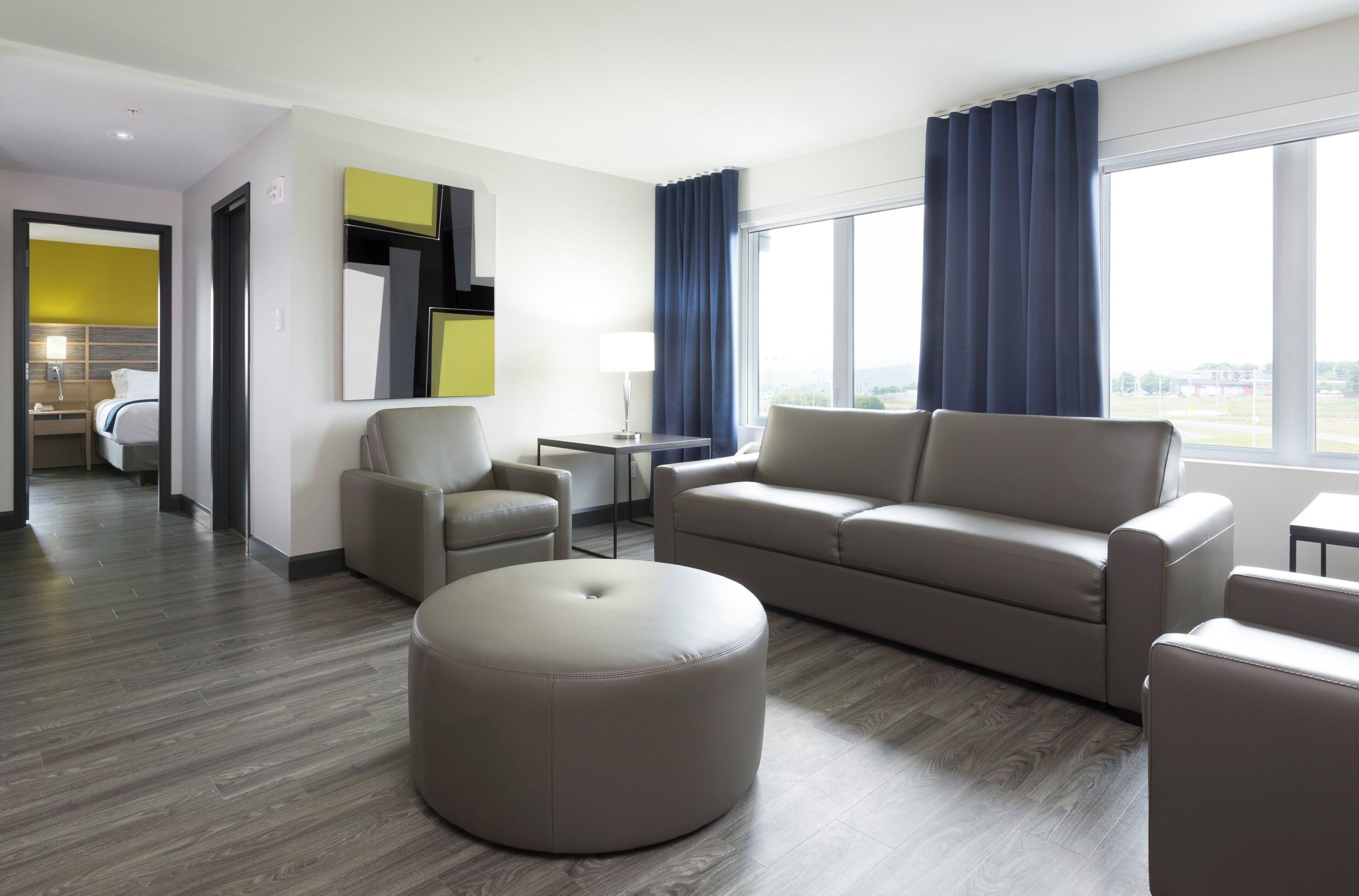 Een moderne hotelkamer met een bank, poef, fauteuils, een klein tafeltje en een lamp. Er is een groot raam met gordijnen en uitzicht op het omliggende landschap.