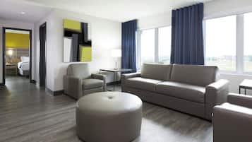 A modern hotel room with a sofa, ottoman, armchairs, a small table, and a lamp. There is a large window with curtains and a view of the outside.