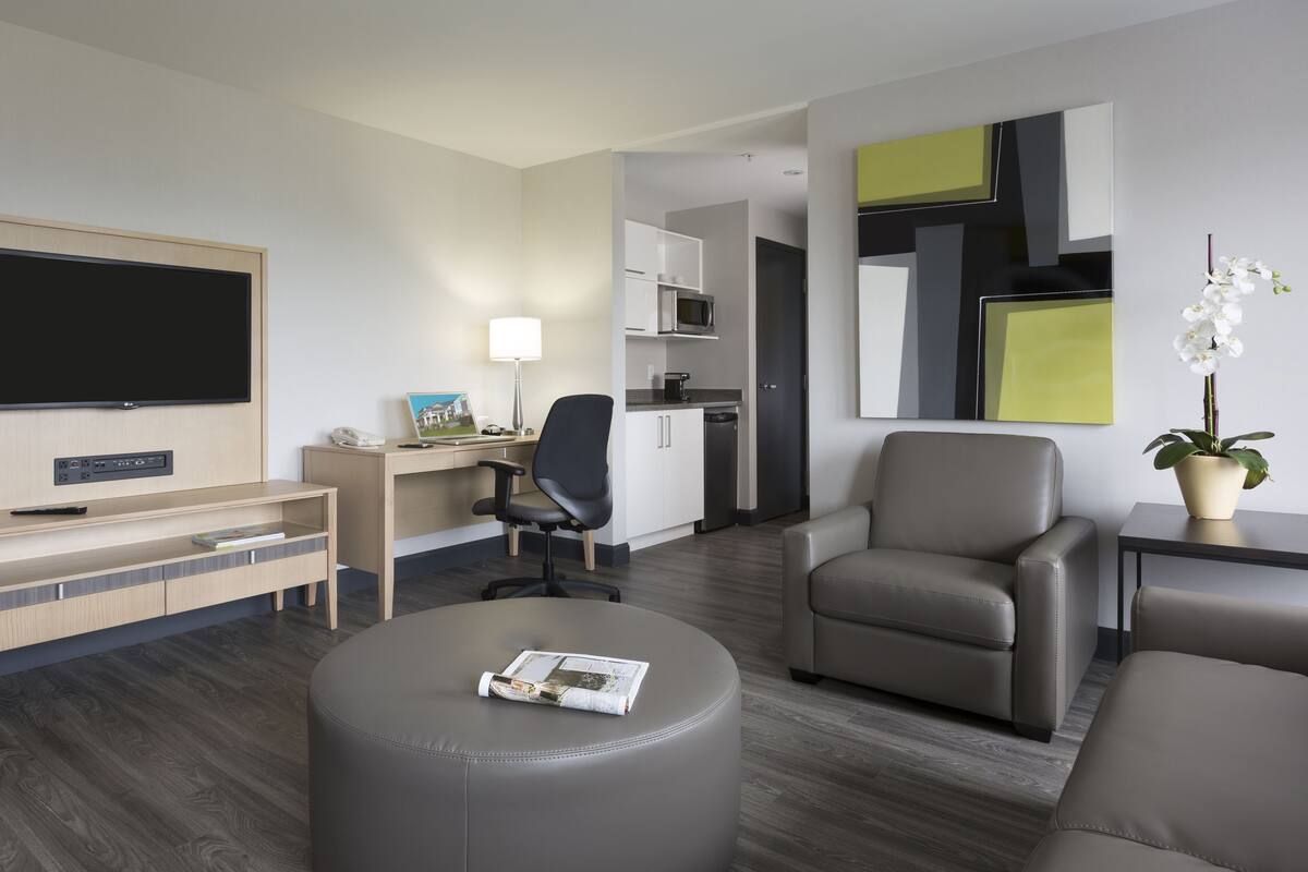 A hotel room with a flat-screen TV, a desk with a lamp, a chair, a sofa, and a small table with a plant.
