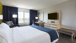 A hotel room with a large bed, a desk, a chair, a TV, and a window with curtains.