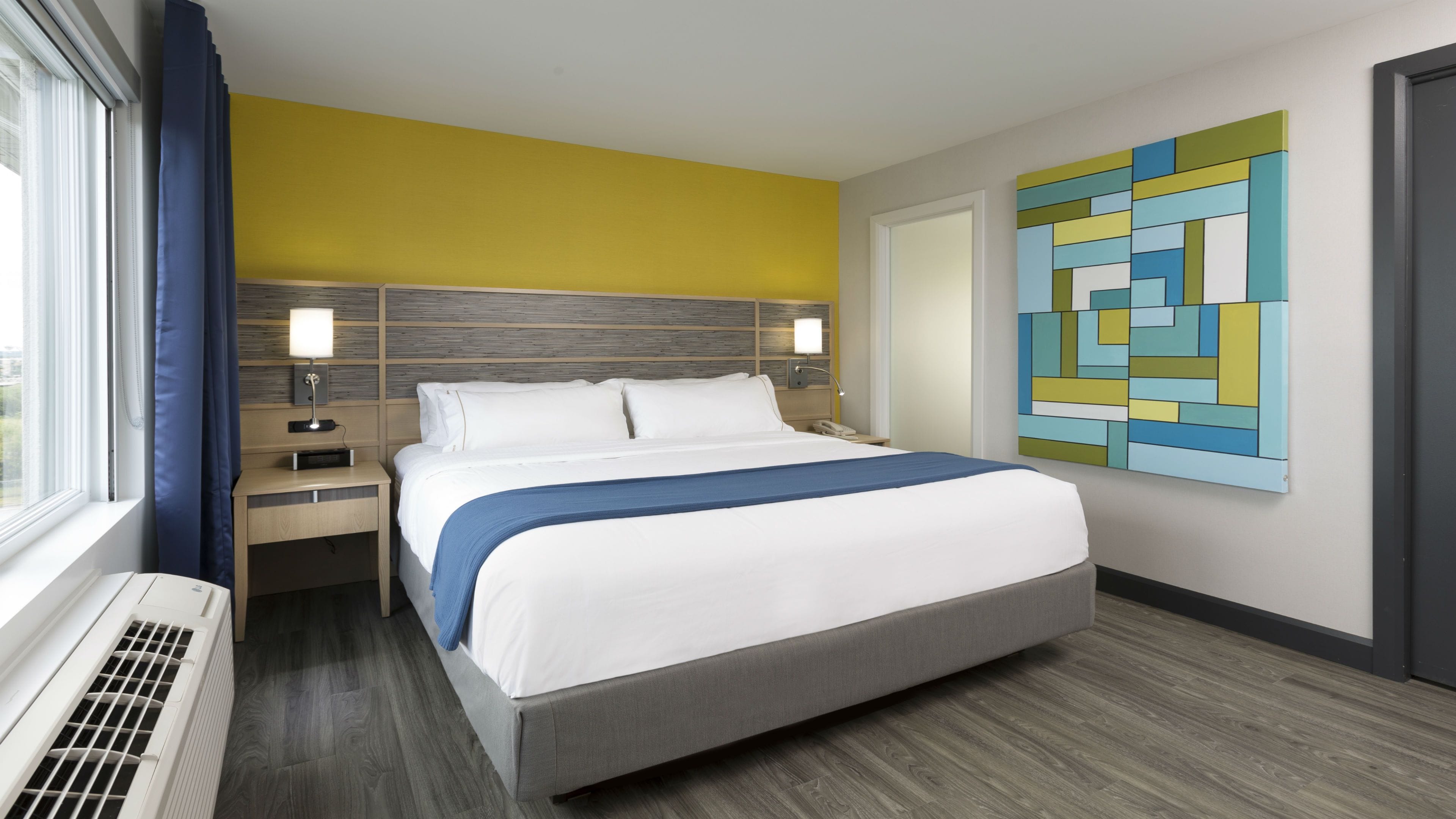 Habitación de hotel con cama, mesitas de noche, lámpara, un cuadro en la pared y ventana con cortinas.