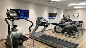 A gym with treadmills, a stationary bike, and a rowing machine. There are two TVs mounted on the wall, one displaying a sports event.