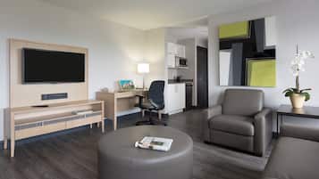 Quarto de hotel com televisão de tela plana, escrivaninha com luminária, cadeira, sofá e mesa pequena com planta em vaso.