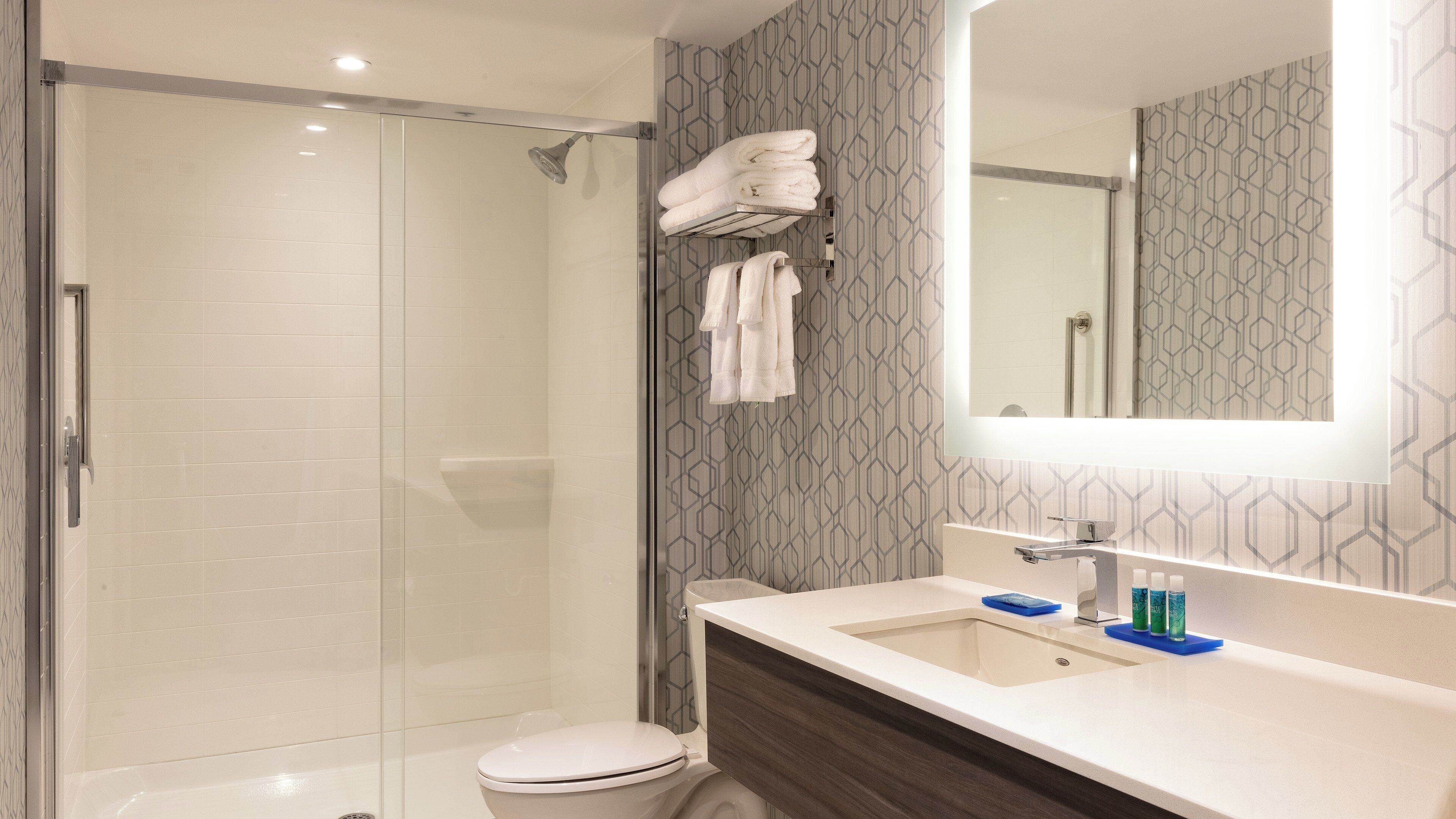 Un baño moderno con ducha envuelta en vidrio, inodoro blanco, tocador con espejo y pared con diseño.