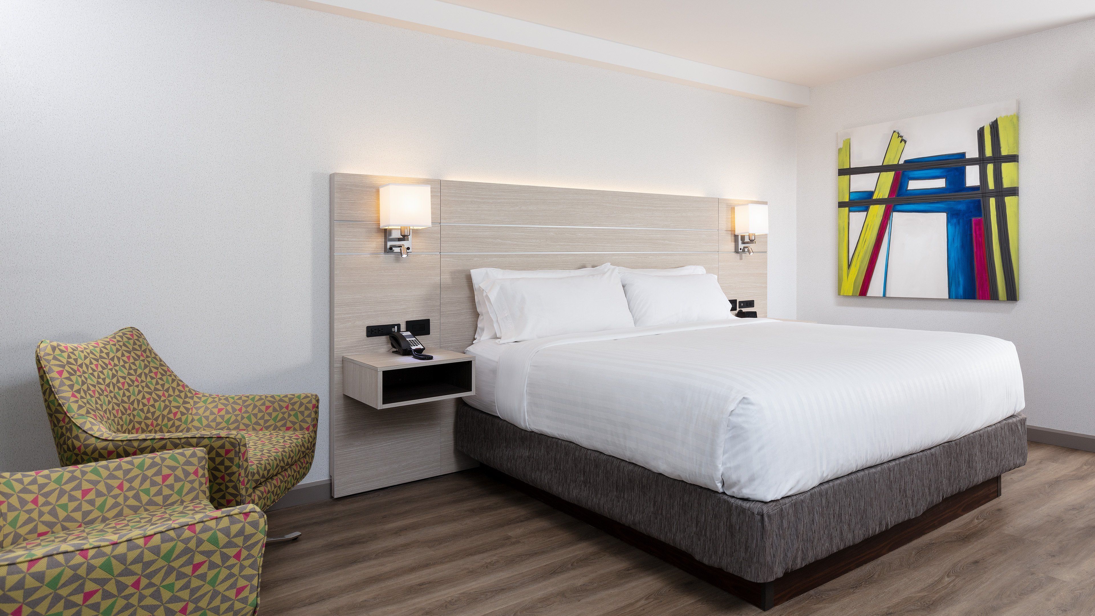 Una habitación de hotel con una cama grande, una mesita de noche con teléfono, una silla y una obra de arte colgada en la pared.
