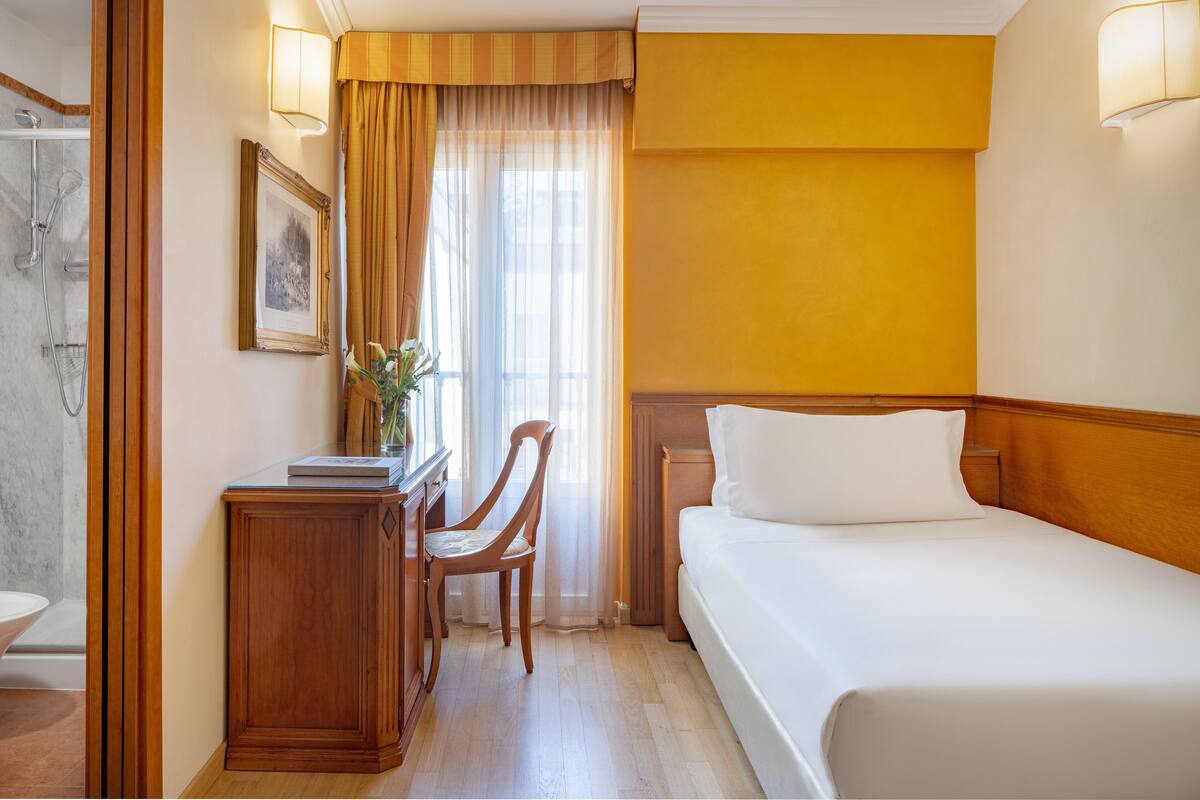 Quarto de hotel com cama, escrivaninha, cadeira, chuveiro e janela com cortinas.