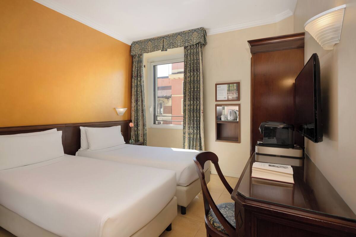 Quarto de hotel com duas camas, uma escrivaninha com cadeira, televisão de tela plana, cafeteira e janela com cortinas.