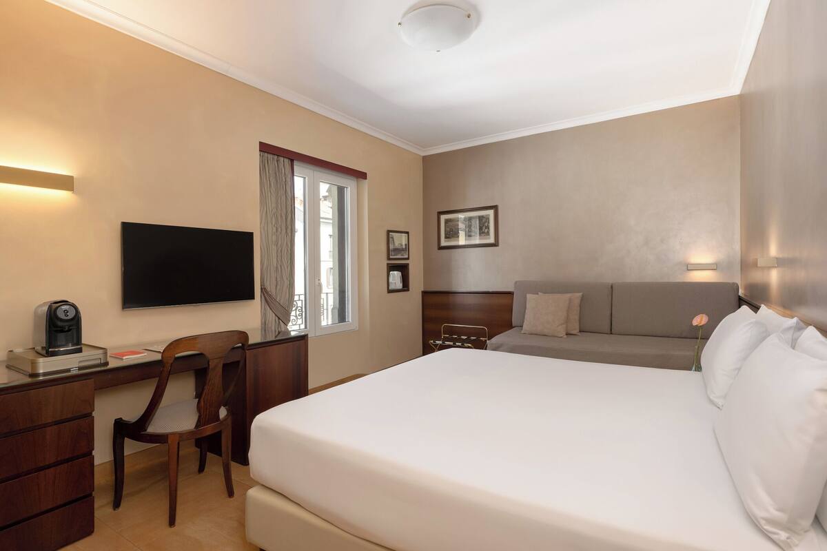 Quarto de hotel com cama, escrivaninha, cadeira, televisão e janela com cortinas.