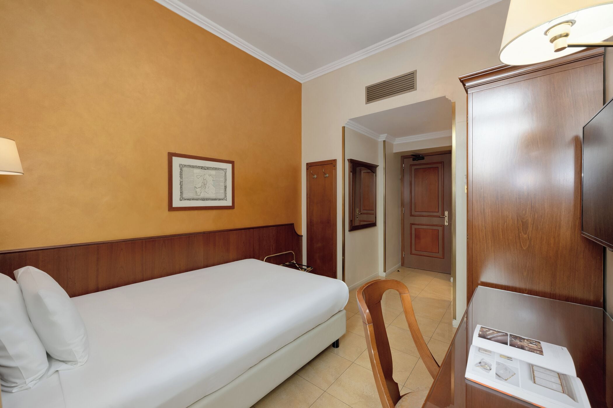 A hotel room with a bed, a wooden chair, a desk with a lamp, and a framed picture on the wall.