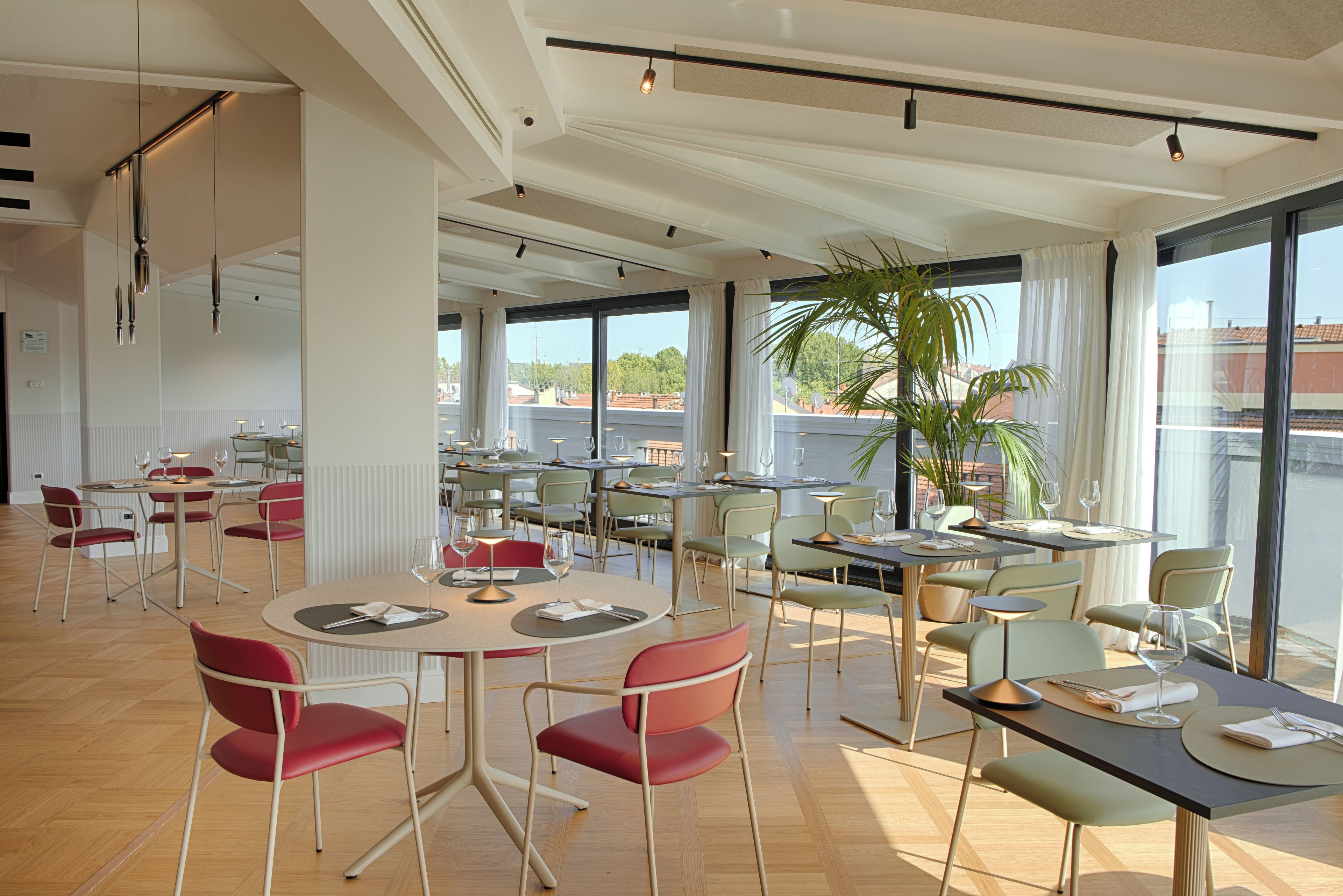 A modern dining area with multiple tables and chairs, large windows with curtains, and a view of buildings outside.