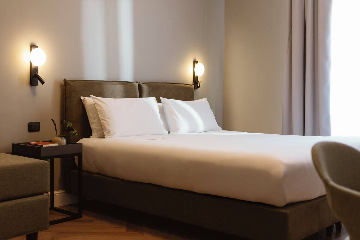 Quarto de hotel com cama, duas mesas de cabeceira, uma cadeira e luminárias fixadas na parede.