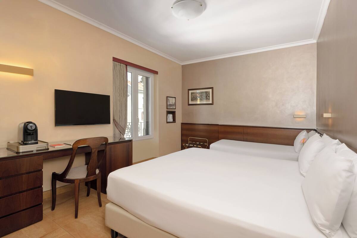 Quarto de hotel com cama, escrivaninha, cadeira, televisão e janela com cortinas.