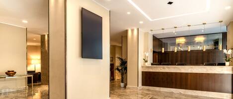 A modern lobby with a reception desk, a large wall-mounted TV, and a potted plant.