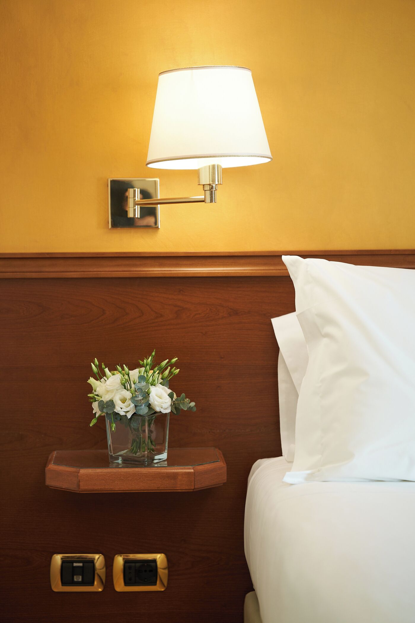 A hotel room with a bed, a bedside lamp, a nightstand with a vase of flowers, and electrical outlets.