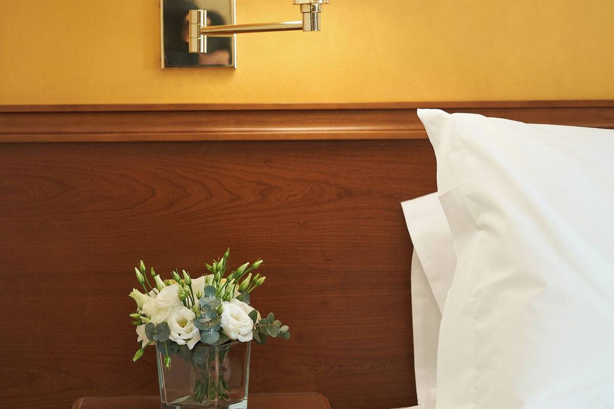 Quarto de hotel com cama, luminária de cabeceira, mesa de cabeceira com um vaso de flores e tomadas elétricas.