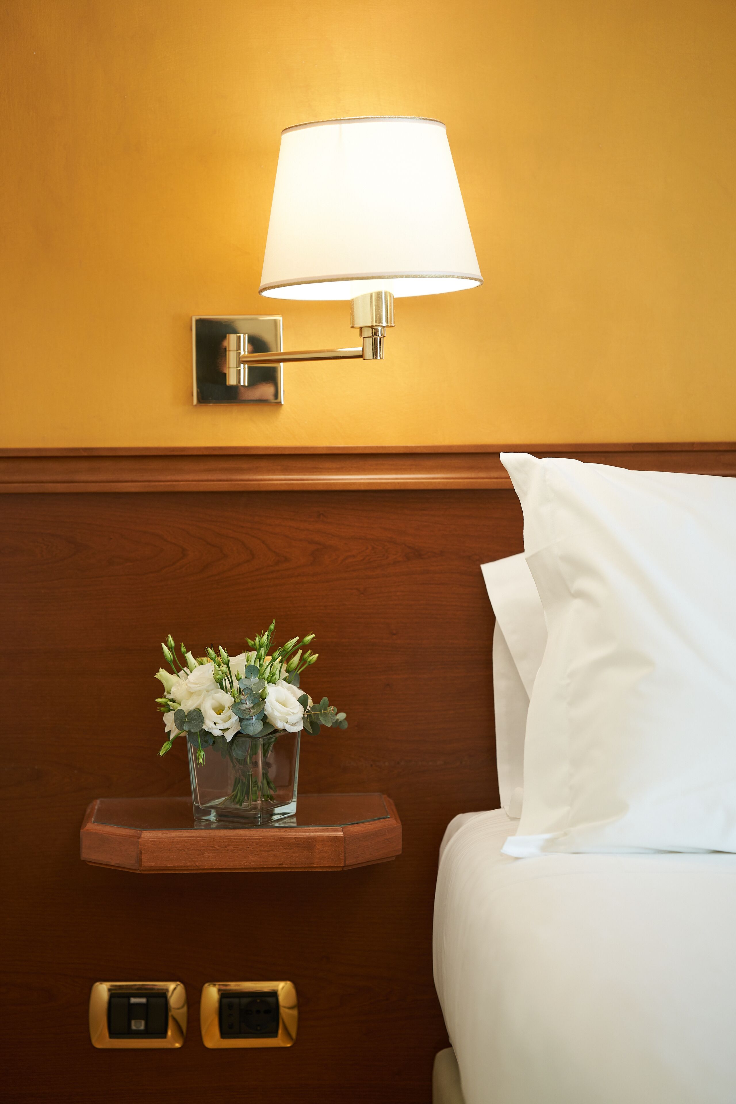 A hotel room with a bedside lamp, a small table with a vase of flowers, and a bed with white linens.