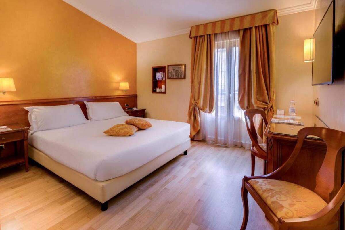 Quarto de hotel com uma cama, mesas de cabeceira de madeira, uma escrivaninha com cadeira, televisão e janela com cortinas.
