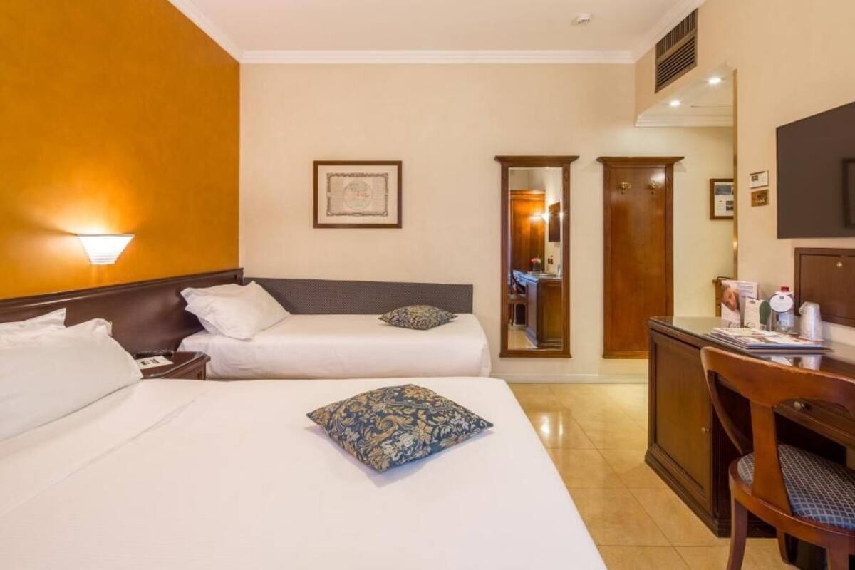 Quarto de hotel com duas camas, uma escrivaninha, uma cadeira, um espelho e uma televisão na parede.