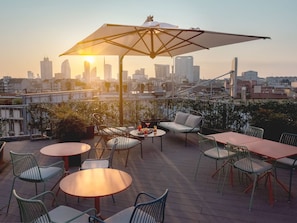 A rooftop terrace with outdoor seating, including round tables and chairs, and a large umbrella providing shade.