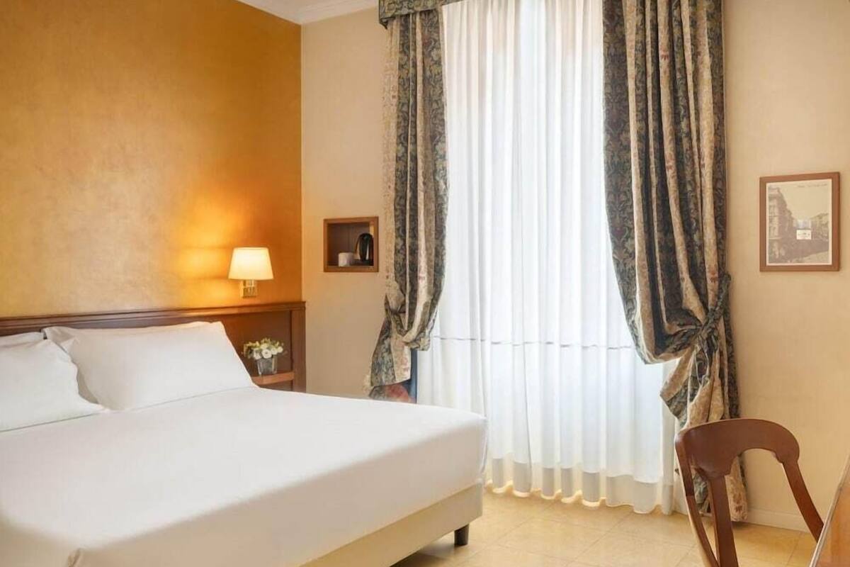 Quarto de hotel com uma cama grande, uma mesinha de cabeceira com abajur, uma cadeira e cortinas.