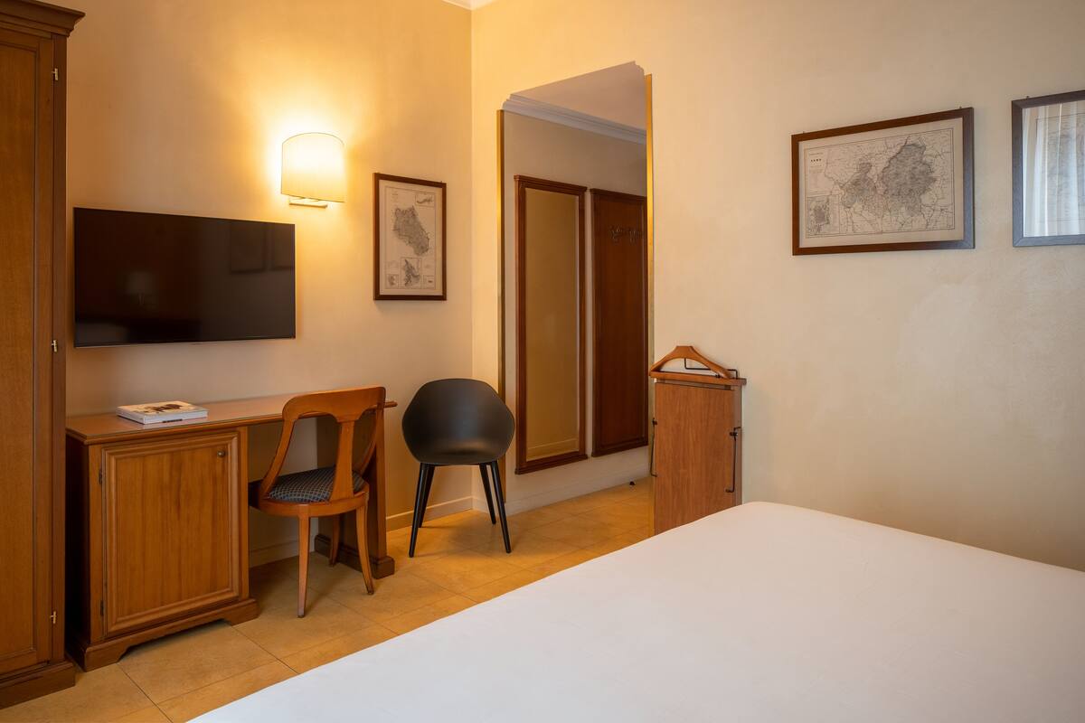 Quarto de hotel com uma cama, uma escrivaninha com cadeira, televisão e quadros nas paredes.