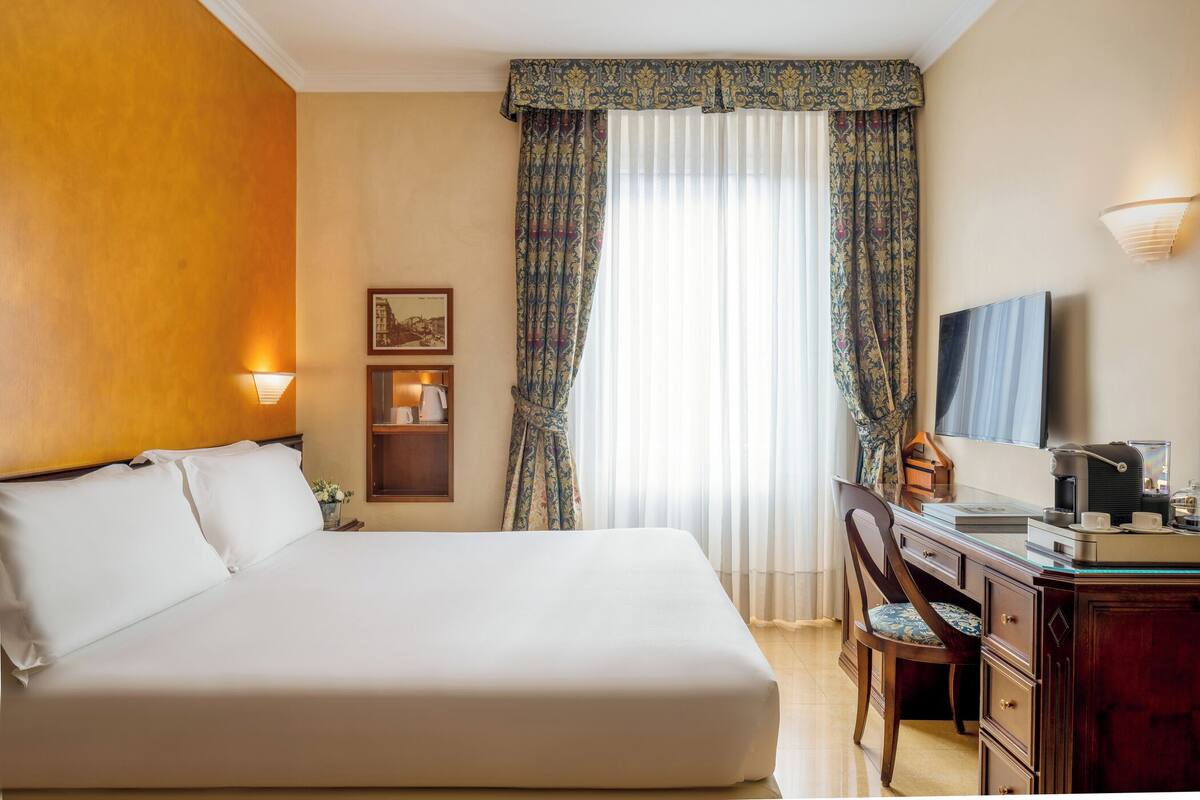 Quarto de hotel com uma cama grande, uma escrivaninha com cadeira, televisão e luminária.