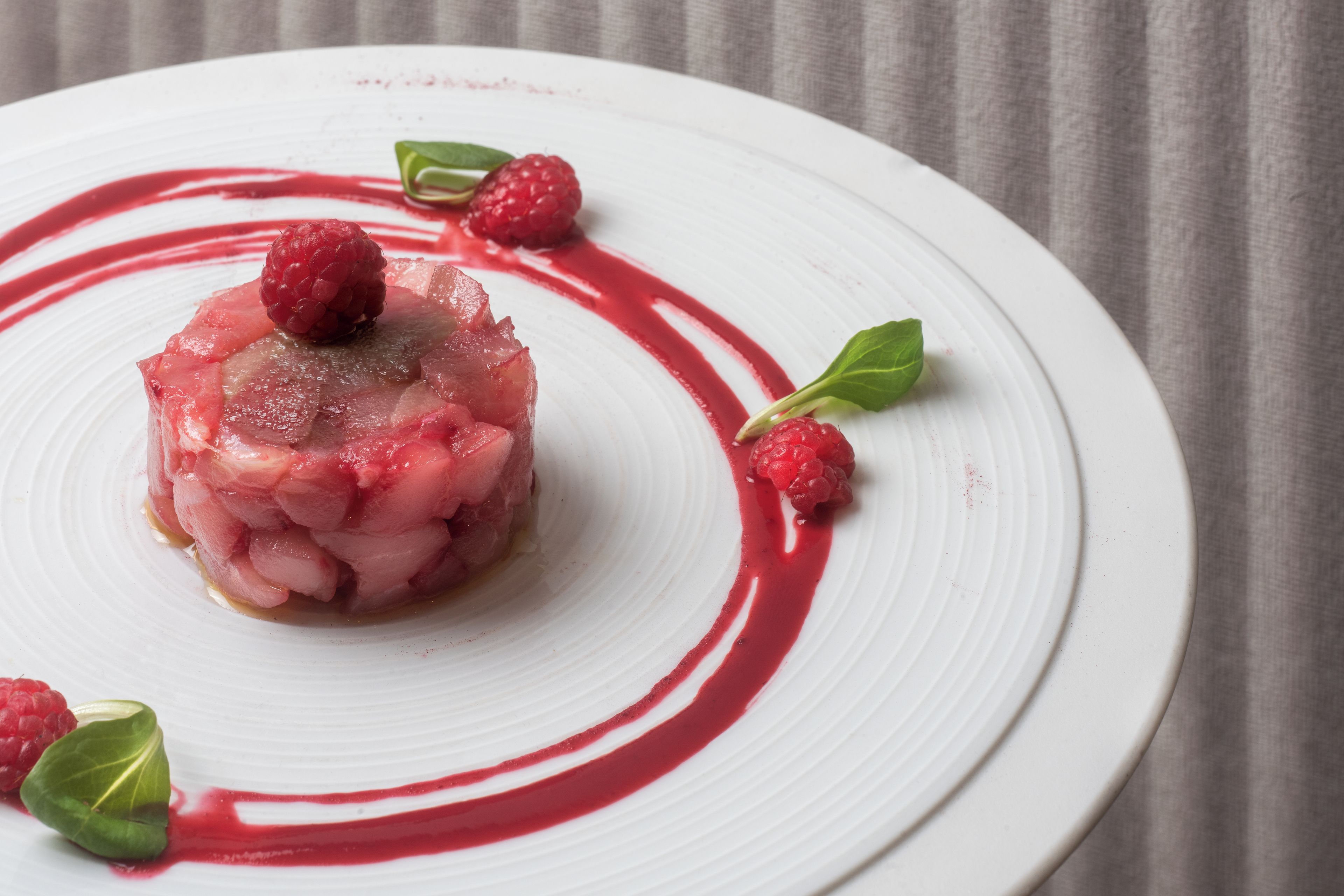 A white plate with a central food item, surrounded by raspberry leaves and a red sauce.