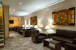 Un'ampia hall con pavimento in marmo, divani e poltrone in pelle, con un grande quadro decorativo e una scala a sinistra.