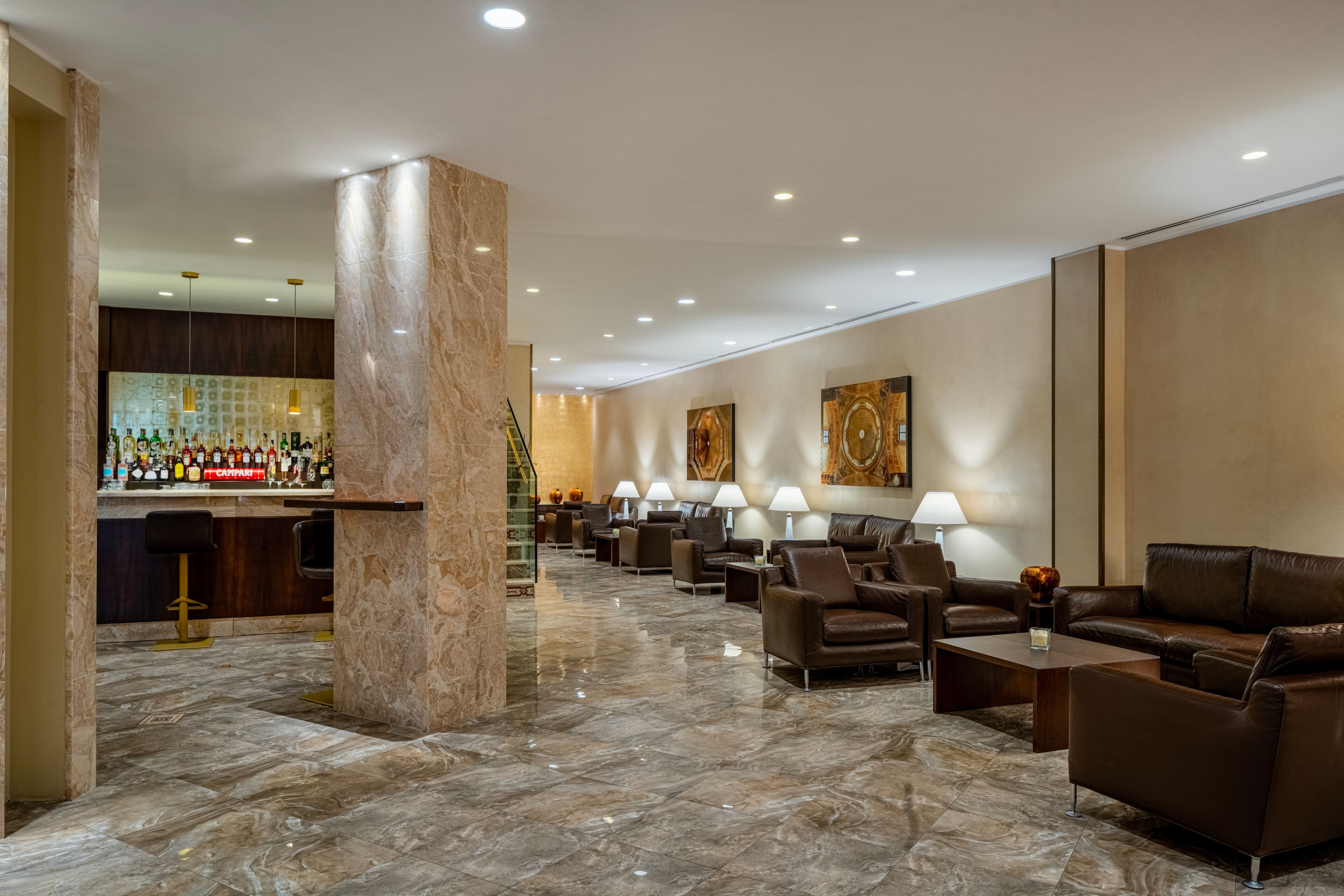 A spacious lobby with a marble floor, leather sofas, a bar area with various liquor bottles, and a staircase leading to an upper level.