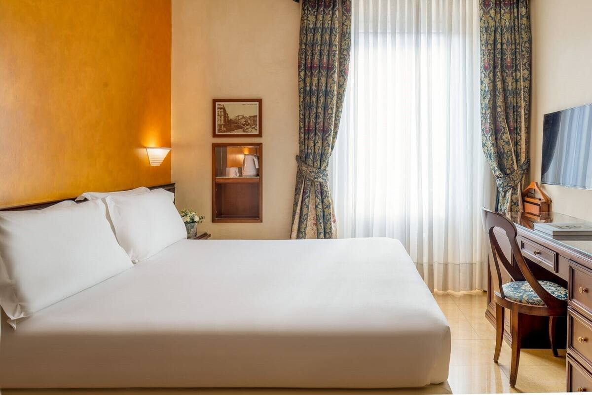 Quarto de hotel com uma cama grande, uma escrivaninha com cadeira, um quadro na parede e uma janela com cortinas.
