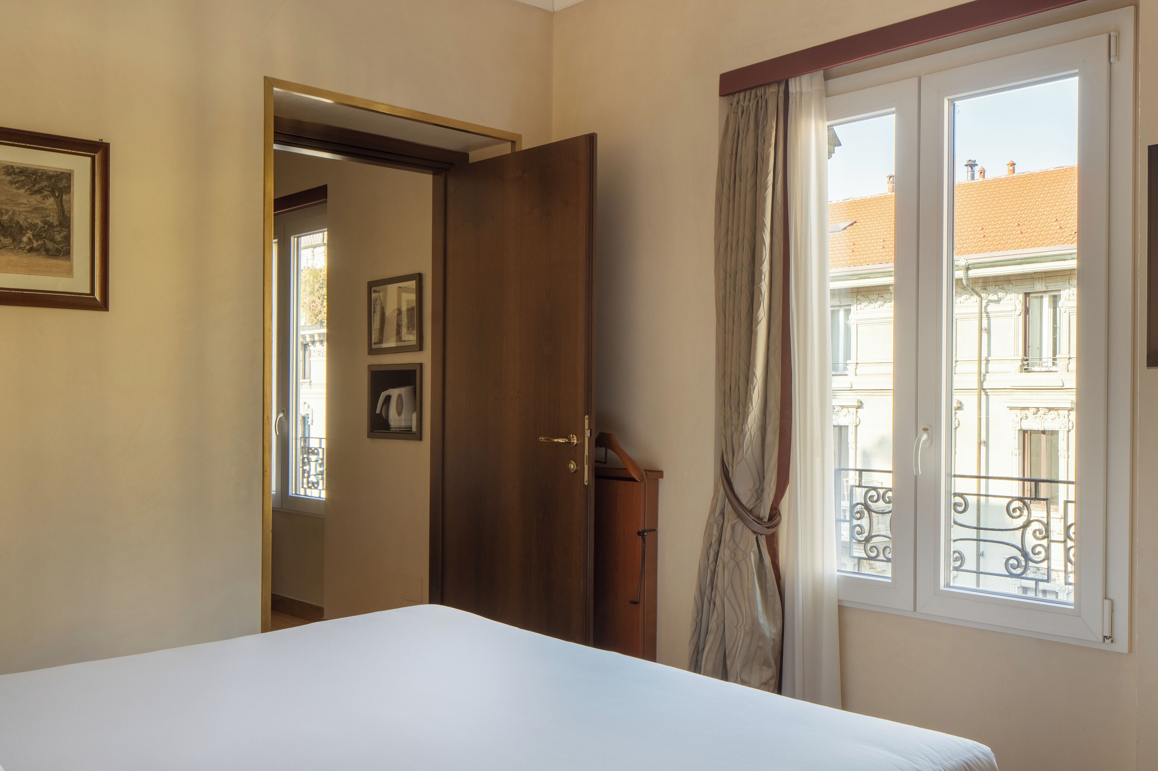 A bedroom with a bed, a wooden door, a window with a view of buildings, and a curtain.