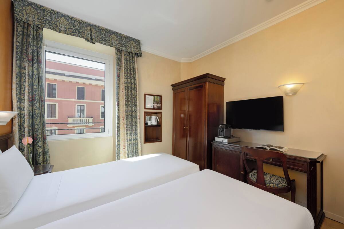 Quarto de hotel com duas camas, televisão, escrivaninha com cadeira, janela com cortinas e vista para um prédio.