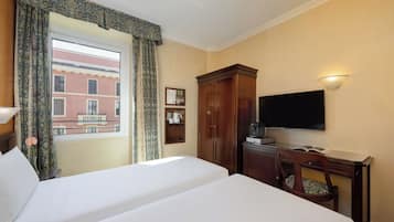 A hotel room with two beds, a television, a desk with a chair, a window with curtains, and a view of a building outside.