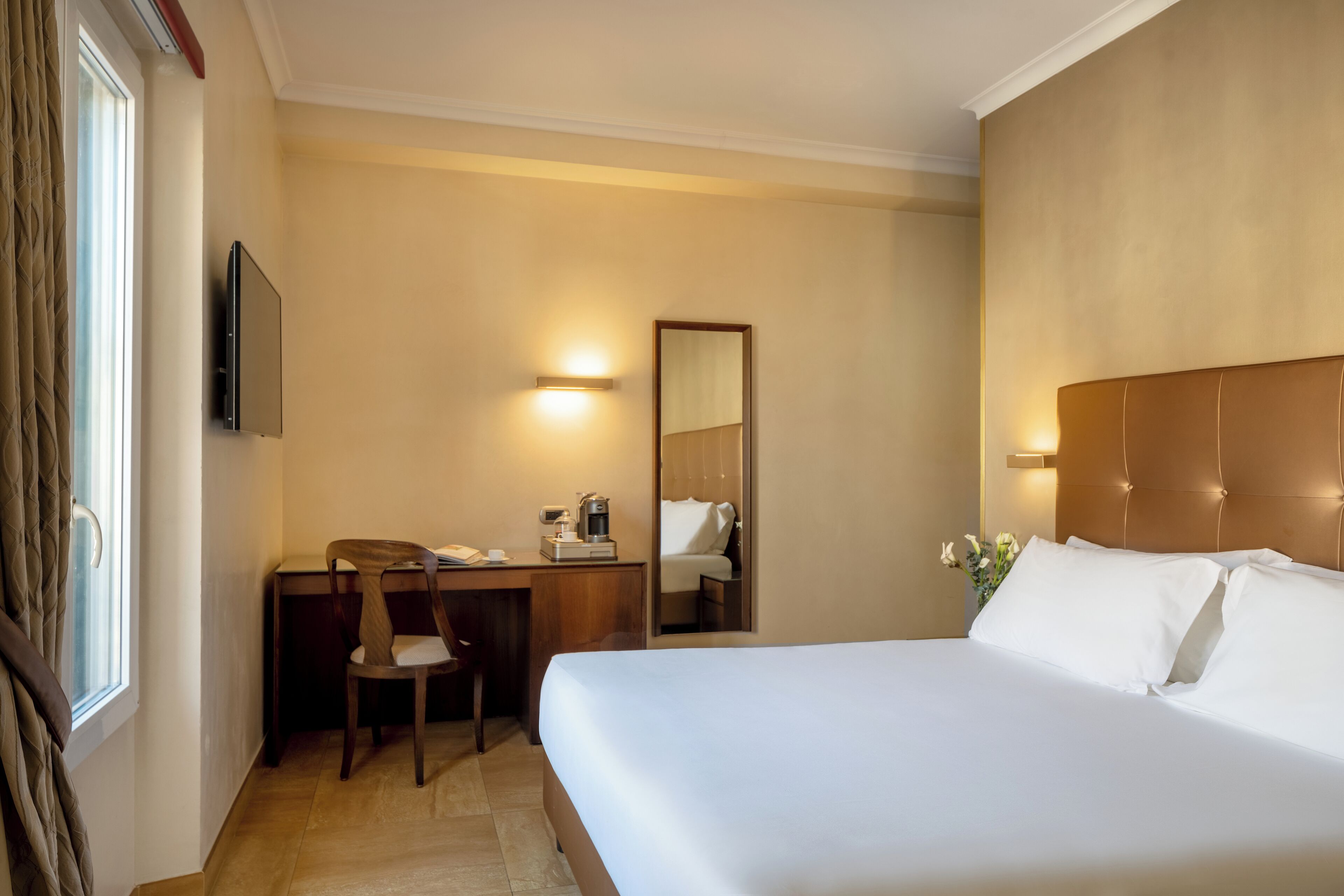 Habitación de hotel con cama, escritorio con silla, cafetera, televisor y espejo.