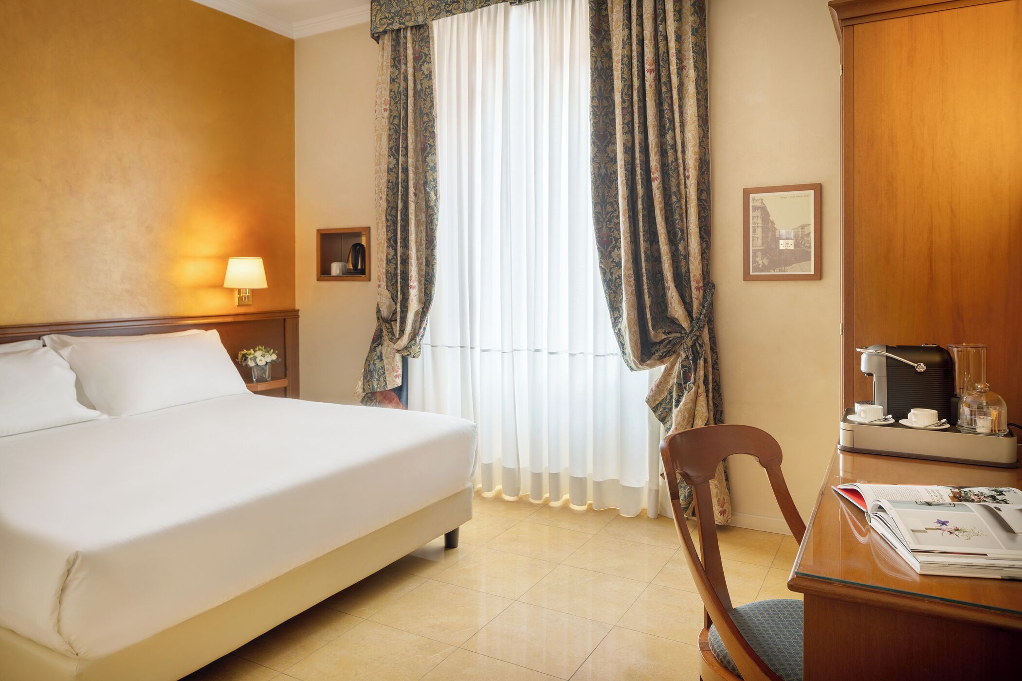 A hotel room with a large bed, a desk with a coffee maker, a chair, and curtains.