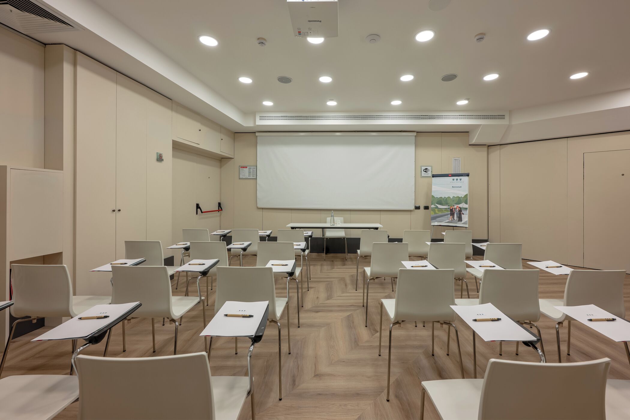 A conference room with rows of white chairs, a large projection screen, and a whiteboard.
