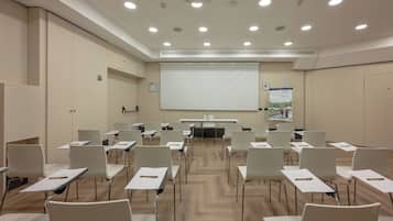 Une salle de conférence avec des rangées de chaises blanches, un grand écran de projection et un tableau blanc.