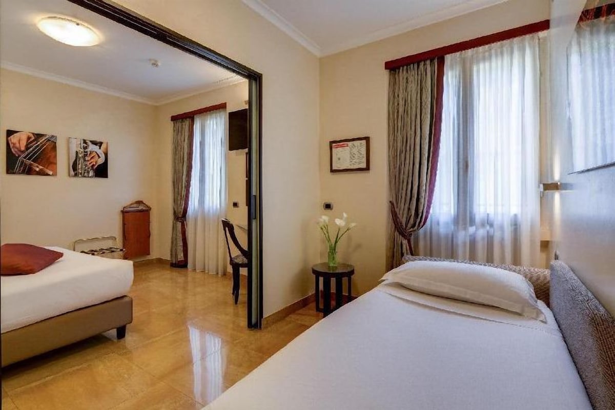 Camera d'albergo con due letti, uno specchio, un tavolino con un vaso e un quadro appeso al muro.