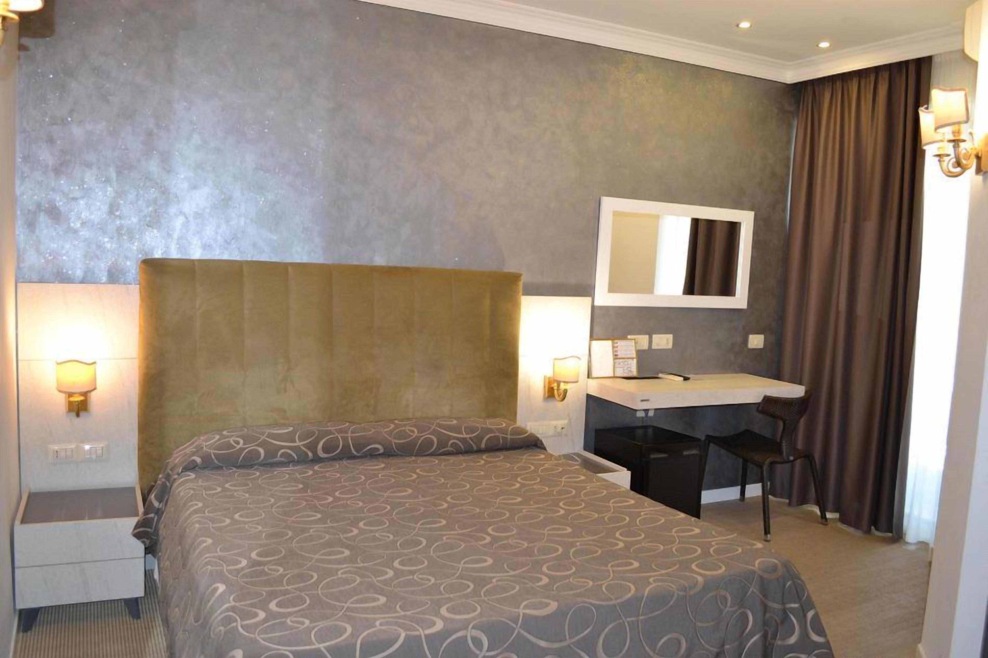 A hotel room with a large bed, a desk, a chair, a mirror, and two wall-mounted lamps.